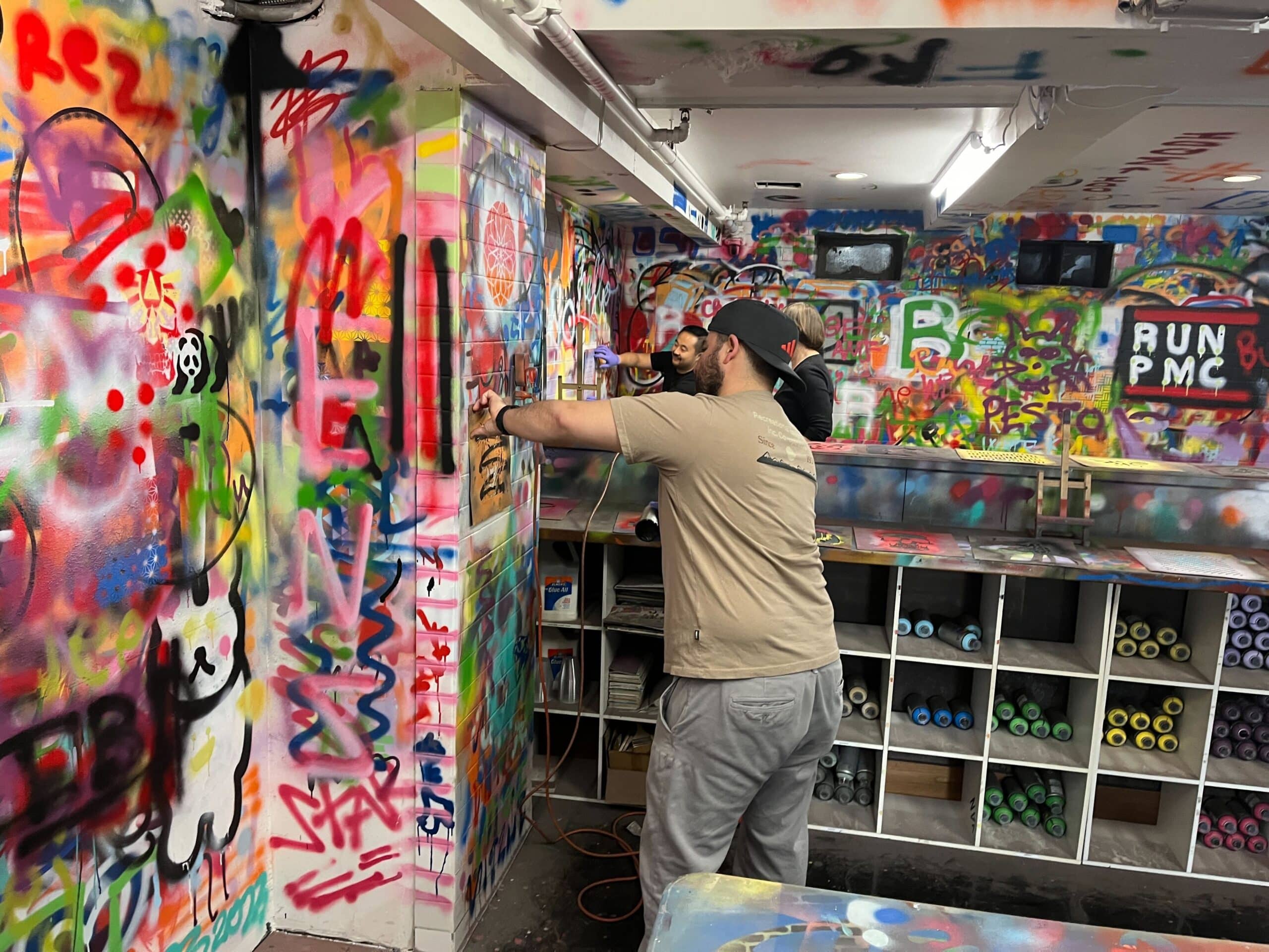 A person in a beige t-shirt and grey pants is spray-painting a colorful graffiti mural in a brightly lit studio. The walls are covered in vibrant, layered graffiti art, and shelves filled with cans of spray paint are visible in the background.