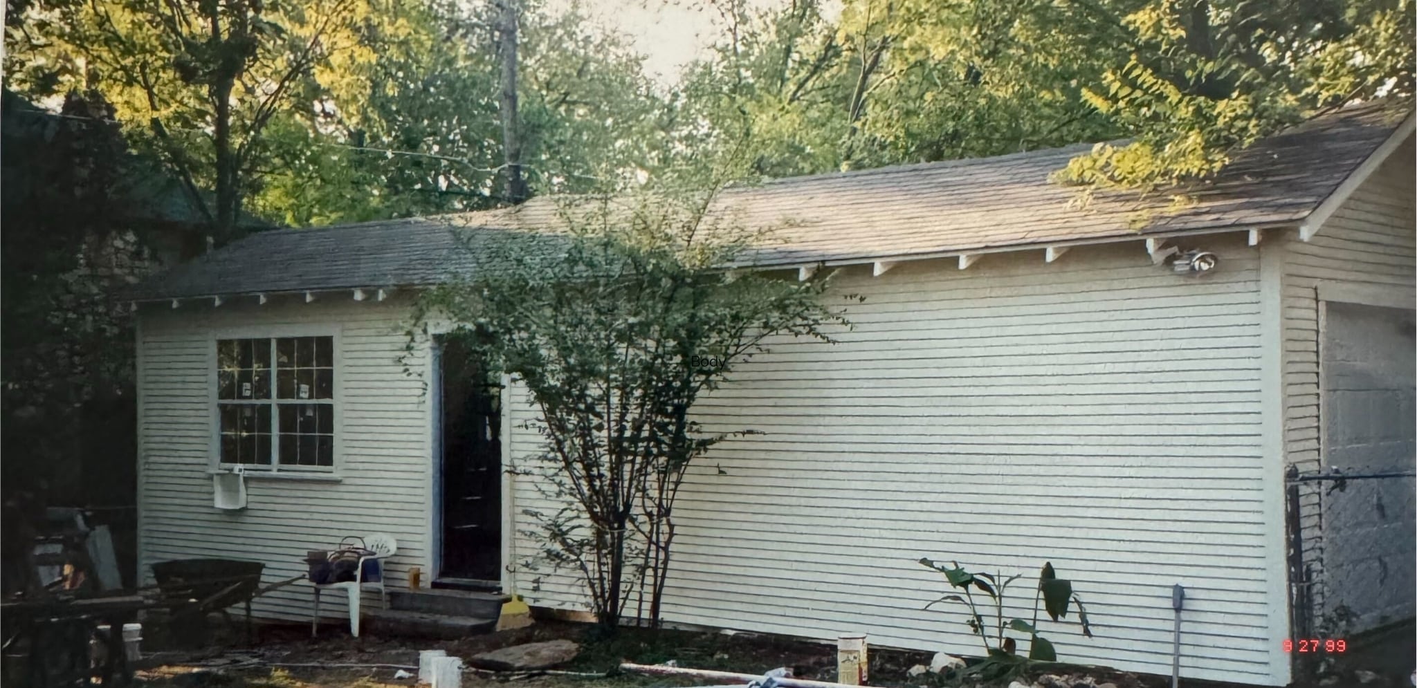 An off white residential garage building with an open side door. One tree grows next to the door. The scene is sunny with wisps of light filtering through trees behind the building. A date stamp from a camera reads 9.27.99.