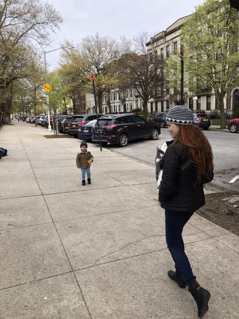 A woman with long hair and a striped hat walks on a city sidewalk, holding a baby. A small child in a brown coat stands several feet ahead. Trees and parked cars line the street.