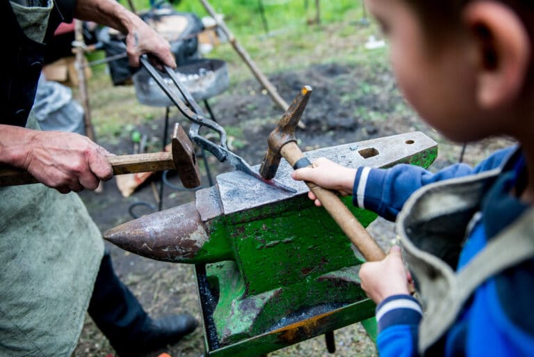 A child and an adult use hammers and tongs to shape hot metal on an anvil outdoors, practicing blacksmithing together—demonstrating authentic craftsmanship with no fake work involved.