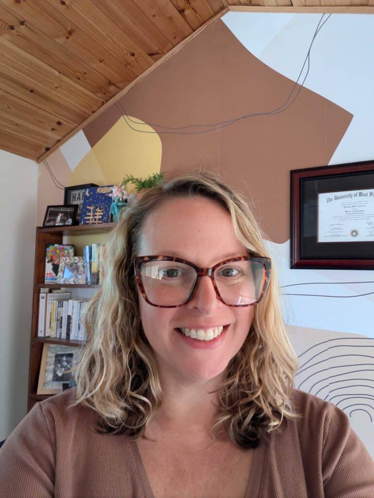 A woman with wavy blonde hair and large glasses smiles in a home office with bookshelves, framed diploma, and geometric wall art behind her.