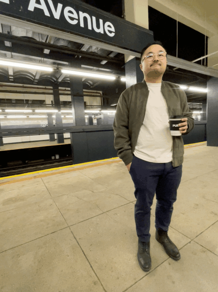 A person stands on a subway platform beneath a sign that reads Avenue, holding a coffee cup and looking ahead. The platform is empty, and the background shows subway tracks and station lights.