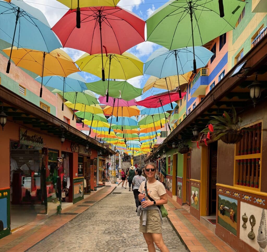 A woman smiles while standing on a colorful street lined with shops and hanging plants, with vibrant umbrellas suspended above, creating a festive atmosphere.