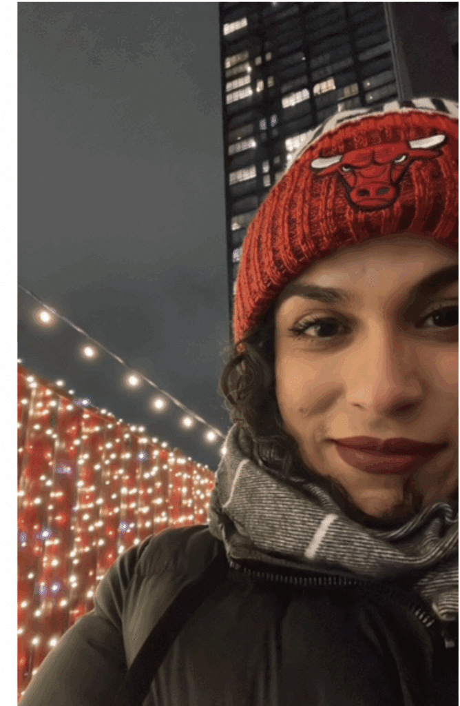 A person wearing a red Chicago Bulls beanie and scarf smiles outdoors at night, with festive string lights and a tall building in the background.