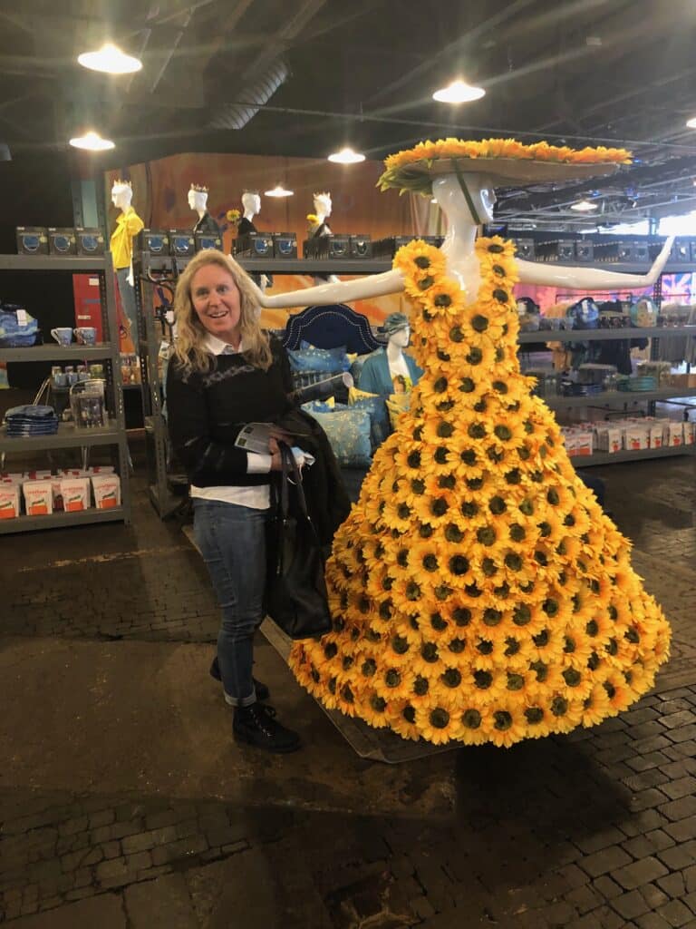 A woman stands smiling next to a mannequin dressed in a gown and hat covered entirely with bright yellow sunflowers, displayed inside a store with various products on the shelves.