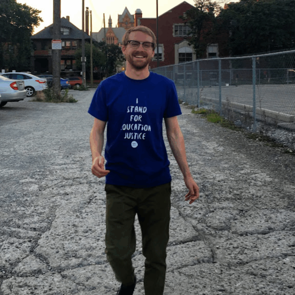 A person with glasses and a beard walks on a gravel lot, smiling. They wear a blue shirt that reads I stand for education justice. Buildings and a fence are in the background.