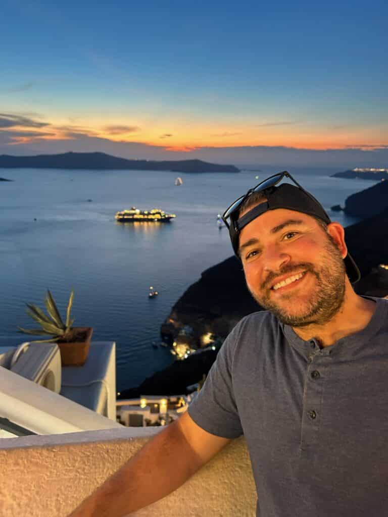A man smiles at sunset on a balcony overlooking the sea, with boats and islands in the background. The sky is colorful, and city lights are visible along the coastline.
