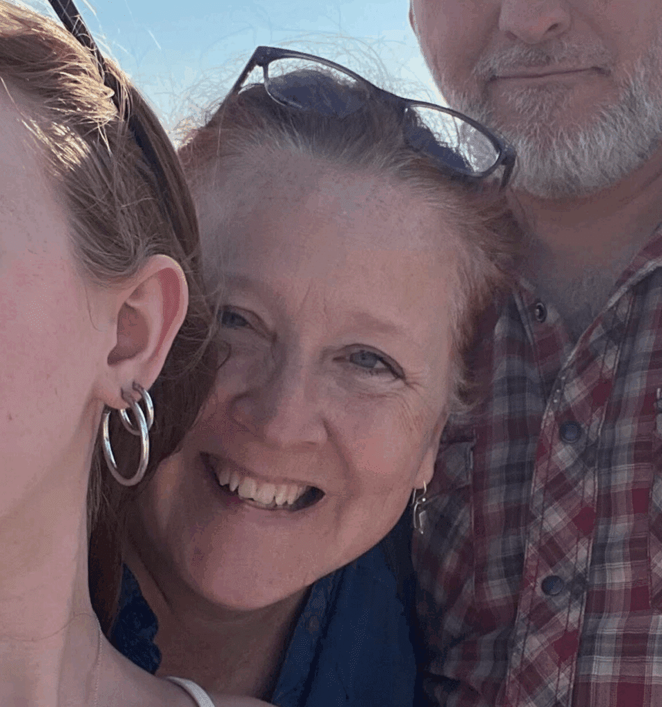 Three people pose closely together, smiling in bright sunlight. Only part of the face of the person on the left is visible. The woman in the middle wears glasses, and the man on the right has a beard and checked shirt.