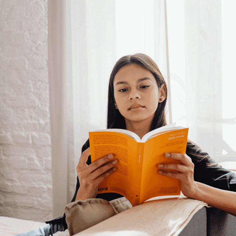 A young girl sits on a couch reading a bright yellow book, with sunlight streaming through sheer curtains behind her.