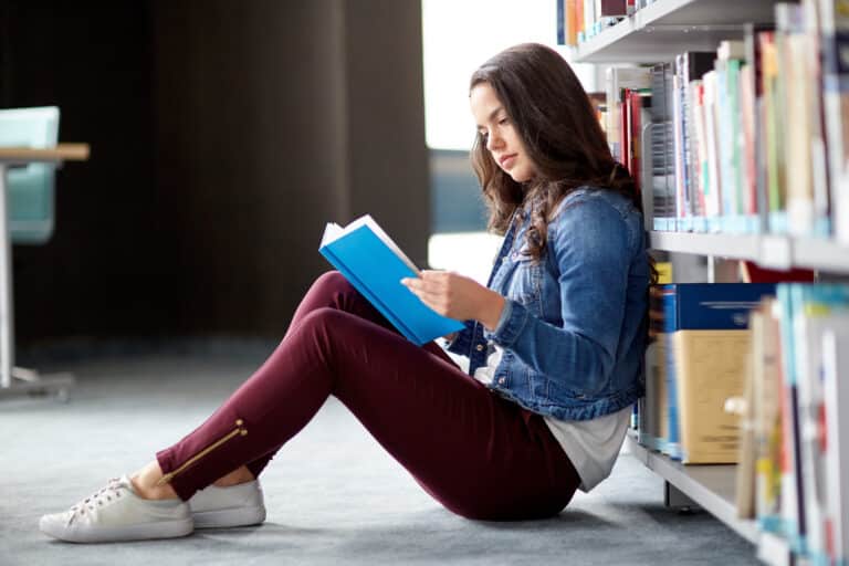 A young woman sits on the library floor, leaning against a bookshelf, immersed in independent reading as she enjoys a blue book. She wears a denim jacket and maroon pants, while sunlight filters in from a nearby window.