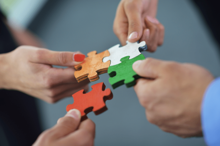 Four hands each hold a different colored puzzle piece, working together to connect them. The puzzle pieces are orange, white, green, and brown—symbolizing the importance of instructional material coherence as each piece fits seamlessly into the whole. The background is blurred.