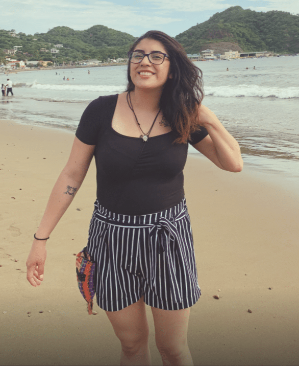 A woman with glasses smiles while standing on a sandy beach, wearing a black top and striped shorts. There are waves, people, and green hills in the background under a cloudy sky.