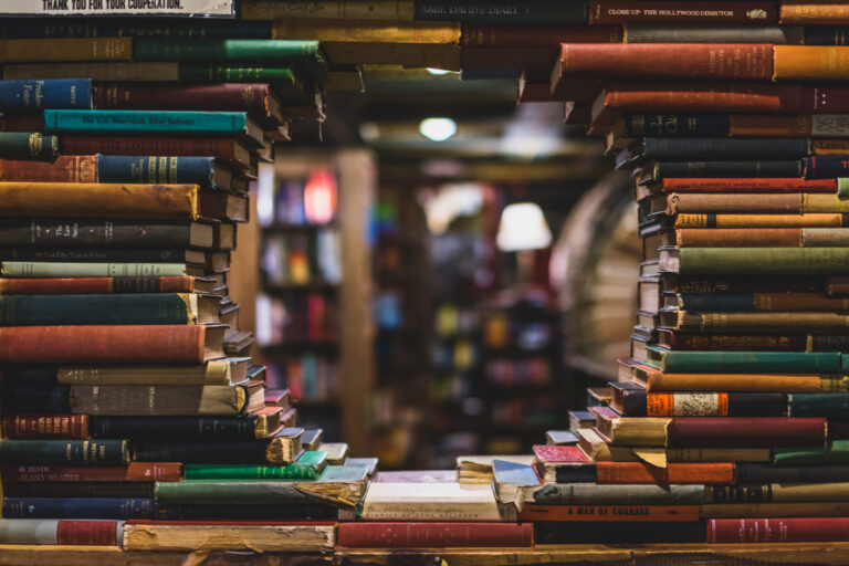 A large stack of colorful, old books forms a window-like opening, revealing more bookshelves in the background inside a cozy, dimly lit bookstore—a hidden haven for English Language Arts enthusiasts to explore timeless stories and classic literature.