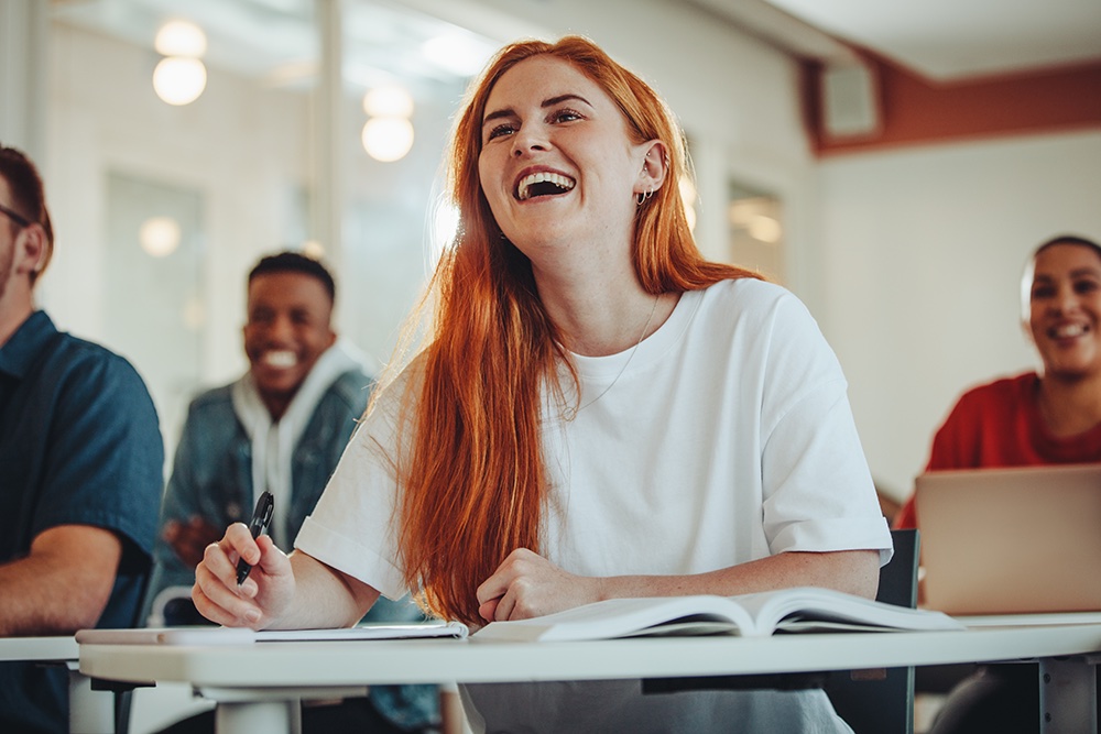 A young woman with long red hair smiles and takes notes in a classroom. She sits at a desk with an open notebook, surrounded by other students, suggesting a positive learning environment.
