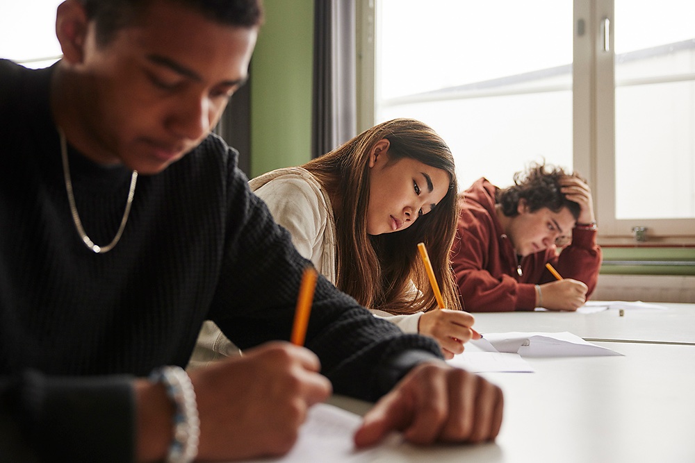 Three students sit at desks, writing on papers. The student in focus appears thoughtful, while another in the background looks stressed, resting a hand on their head. Sunlight comes through a window.