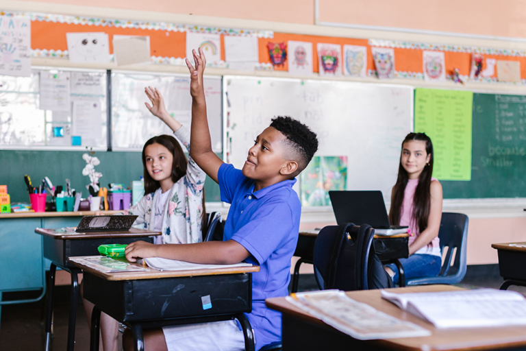 Two students sitting at desks in a classroom raise their hands to answer a question, smiling and looking engaged. Colorful decorations and school supplies are visible in the background.