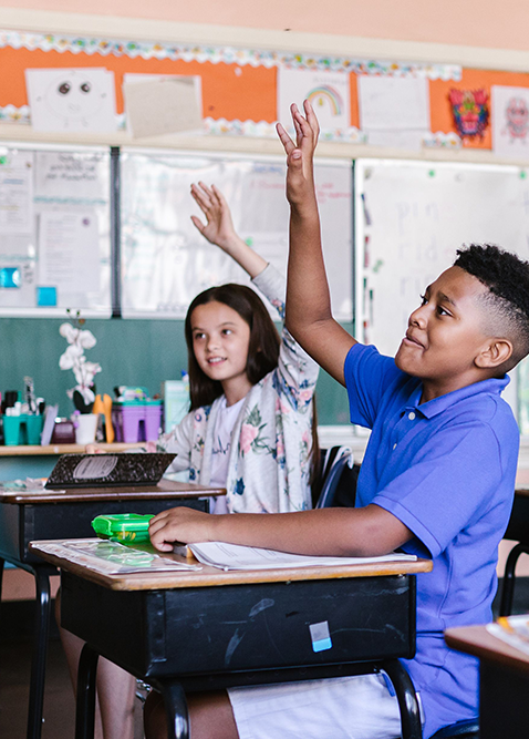 Two students sitting at desks in a classroom raise their hands to answer a question. The classroom walls have papers and drawings displayed. Both children appear engaged and attentive.