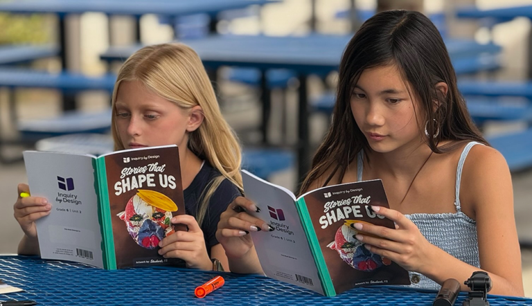 Two girls sit at a blue table outside, reading Stories That Shape Us. One holds a highlighter. Blue benches are visible in the background.