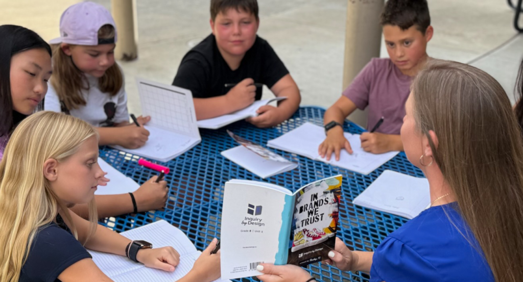 A teacher sits at a blue table outside with five students, all writing in notebooks. The teacher holds a book titled In Brands We Trust. The group appears engaged in a collaborative lesson or discussion.