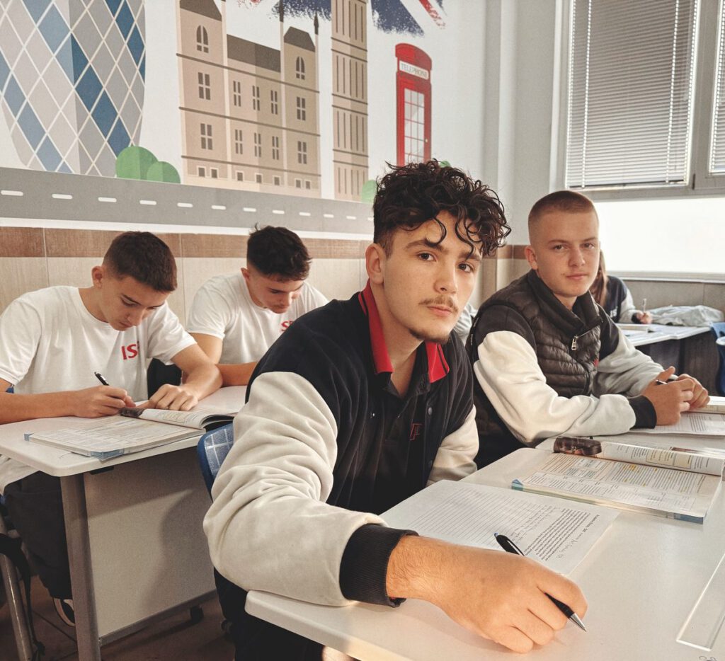 Four male students sit at desks in a classroom, working on worksheets from their literacy curriculum. Two face the camera, while two are focused on their papers. The classroom wall features artwork of buildings, including Big Ben and a red phone booth.