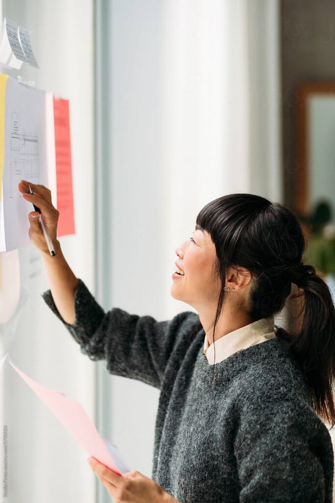 A woman with dark hair in a ponytail smiles while pinning a paper to a bright wall with other documents. She holds more papers in her left hand and appears to be working or organizing information, perhaps arranging materials for a literacy curriculum.