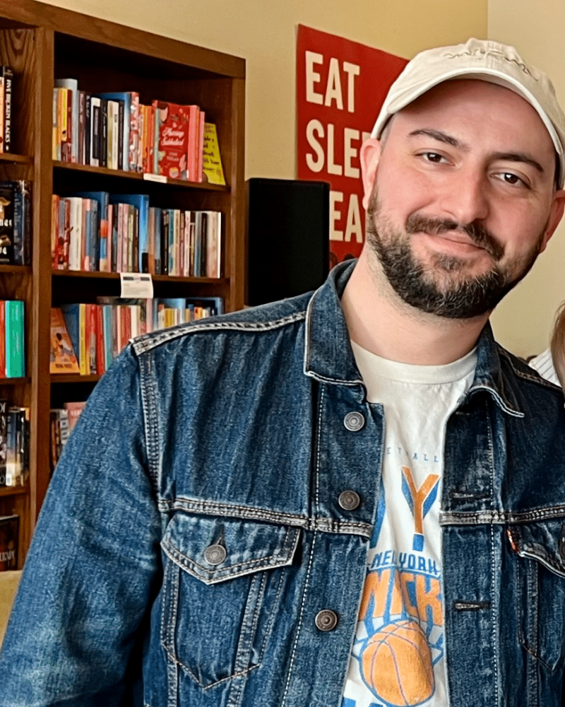 A man with smiling eyes stands in front of a bookshelf full of books.