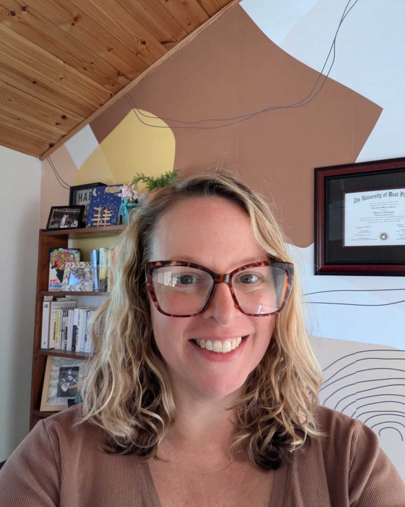 A photograph of a smiling woman standing in her office. A bookshelf behind her is full of books and photos of family and friends.