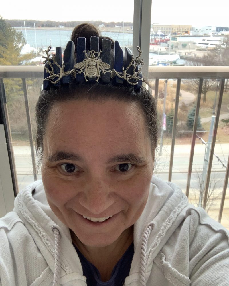 A woman sitting in front of a window wearing a crown with a silver bee on the frtont of it. She is smiling.